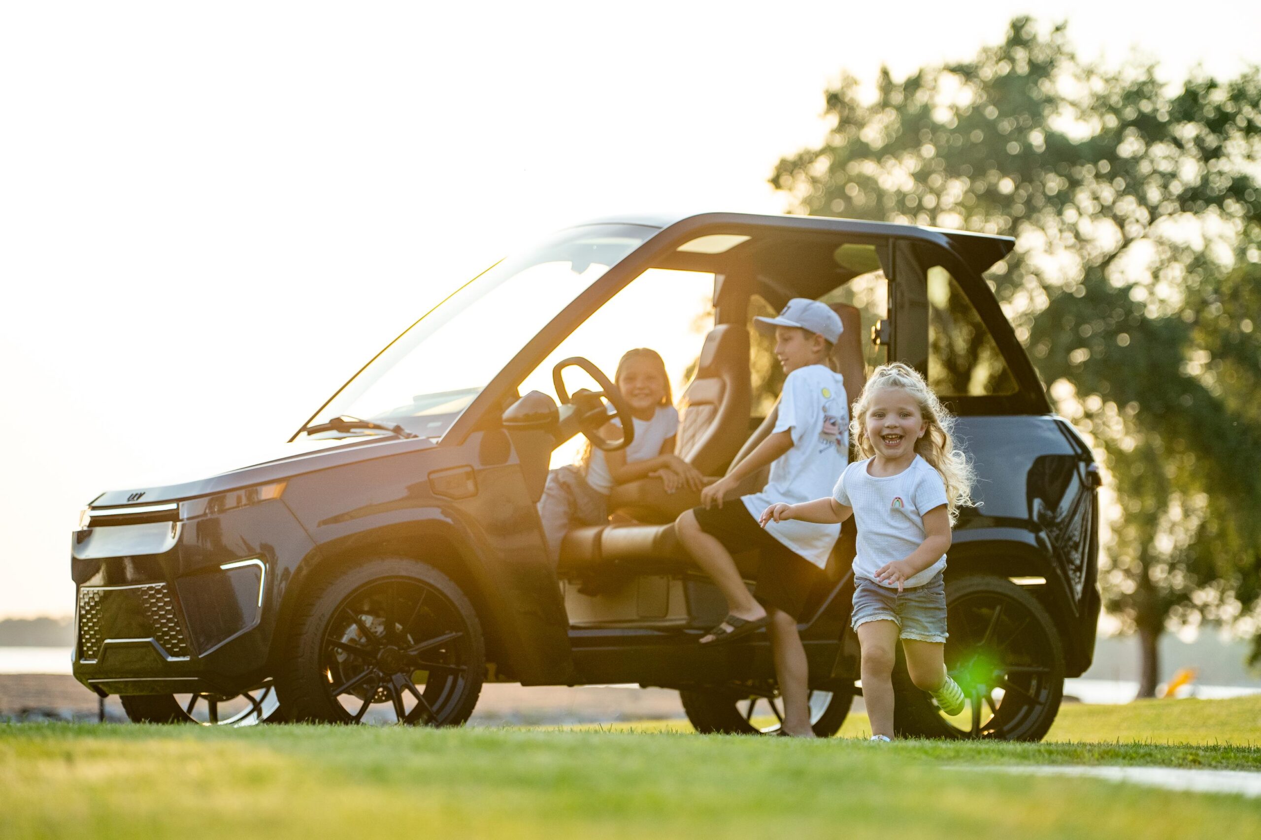 a young girl smiles and runs at the camera as 2 other kids sit inside a ulv model parked beside a lake