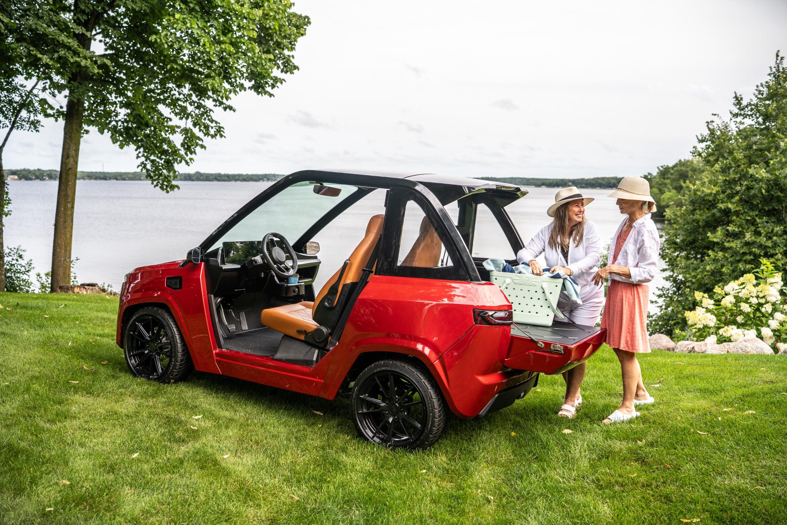 two women unload a picnic basket from the trunk of a red ulv 322 parked next to a lake